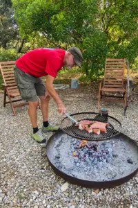 Ein Mann steht am Grillrost der über dem Feuer hängt und wendet das Bratgut.
