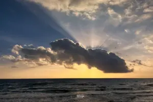Hinter der grossen Wolke leuchtet die Sonne hervor und wirft ihre Strahlen in den Himmel und der Horizont leuchtet golden von der untergehenden Sonne.