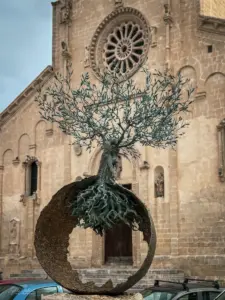 Eine aus bronze gearbeitete Skulptur zeigen eine Olivenbaum mit seinen filigranen Blättern und dem massiven, knorrigen Stamm der seine Wurzeln in der angedeuteten Weltkugel stecken hat.