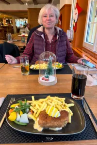 Am Tisch im Restaurant sitzt eine Frau, vor sich den Teller mit Pommes Frites und einem grossen Cordon-Bleu.