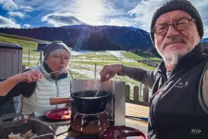 Zwei Personen sitzen draussen auf der Terasse an der Sonne und essen Fondue mit Blick auf verschneiten Kronberg.
