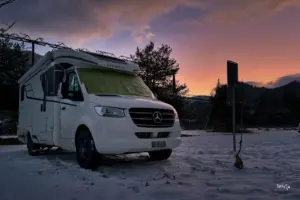 Ein Wohnmobil steht im Schnee auf einem Parkplatz mit einem leuchtenden Sonnenuntergang im Hintergrund.