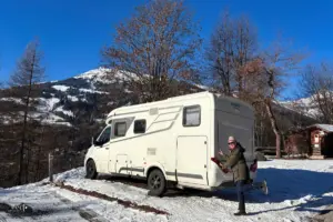 Wohnmobil steht parkiert auf einem Wohnmobilparkplatz im Schnee mit Bergen im Hintergrund.