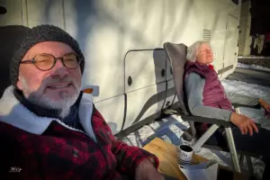 Ein Mann und eine Frau sitzen an der Sonne vor dem Wohnmobil und geniessen im Schnee die Wintersonne.