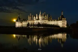 Der aufgehende Mond strahlt hinter dem Schloss Chambord welches sich durch die Beleuchtung vom Nachthimmel abhebt und im Wasser spiegelt.