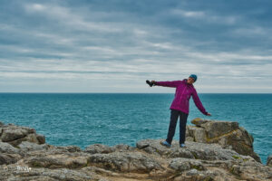 Eine Frau steht auf den Klippen direkt am Meer und trotzt dem Wind.