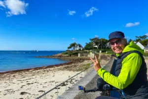Eine Frau mit dem Fahrrad steht an der Küste und winkt in die Kamera.