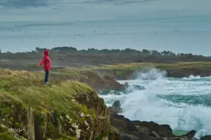 Eine Frau stemmt sich an den Klippen gegen den tosenden Wind und die Wellen klatschen ihre Gist bis über die Klippen hoch.